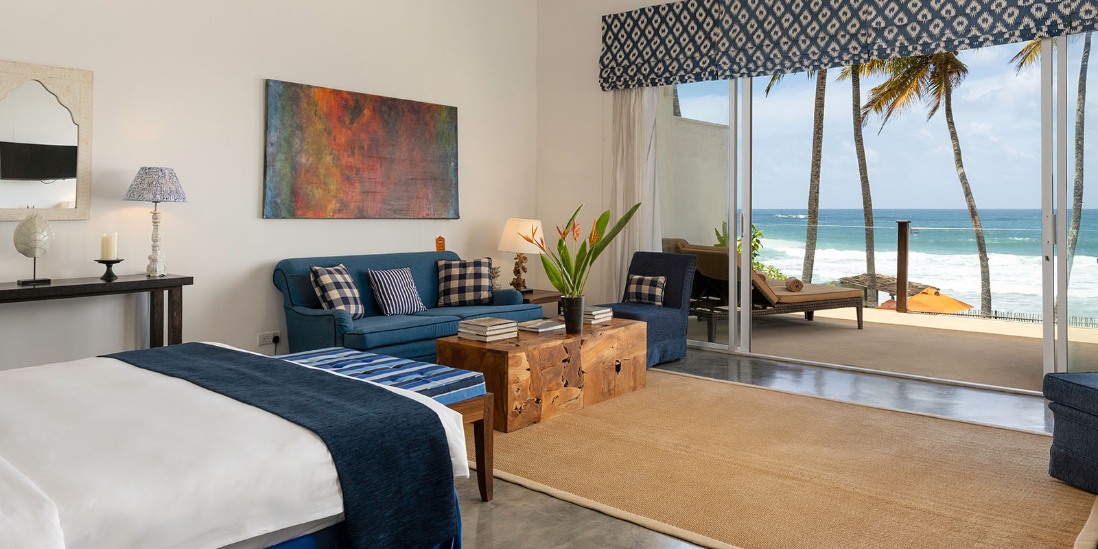 An ocean-facing suite with a white bed, a blue sofa and glass doors open to a balcony over the beach and palms